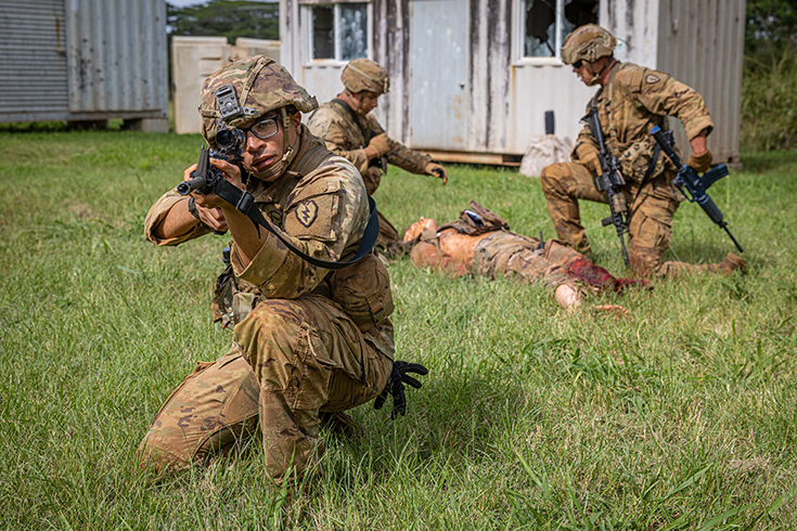 A helmeted, bespectacled Soldier kneels in the grass, pointing his rifle before him. Behind him, two other helmeted Soldiers aid a third Soldier, lying face up on the ground, his left leg seemingly bloodied (though the photograph, in fact, shows a medical training exercise)