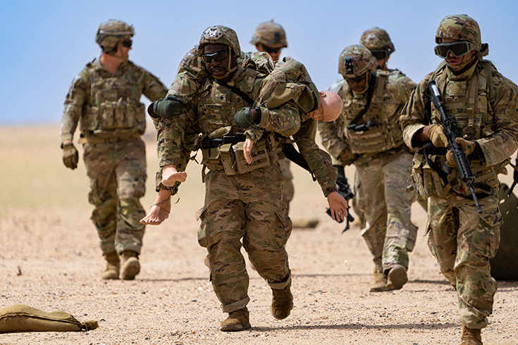 Six helmeted Soldiers wearing vests and sunglasses walk on a sandy plain that stretches to a horizon meeting a cloudless blue sky. The Soldier at the image’s center carries a uniformed dummy on his shoulders.