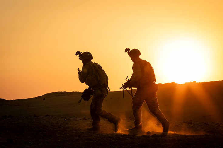 A pair of helmeted Soldiers carrying rifles walk before a ridge, their boots raising dust, as a low sun in a clear sky silhouettes them.