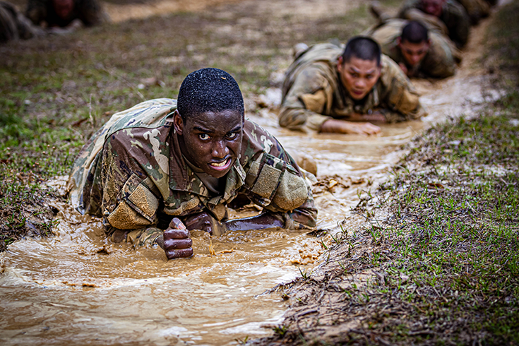 A grimacing Soldier crawls through muddy water, the top of his bare head soiled, and the brown liquid covering his forearms and rising to his chest.