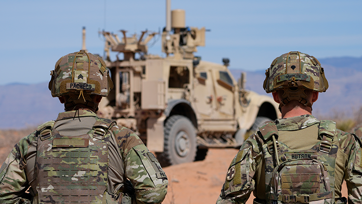Two modern-day Soldiers in helmets and vests stand with their backs to the camera in a sandy, mountainous area near a military vehicle.