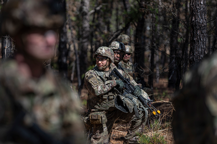 A Soldier in helmet and goggles kneels in a forested area, rifle resting on his knee. A line of three other Soldiers in a similar position is visible at his back.