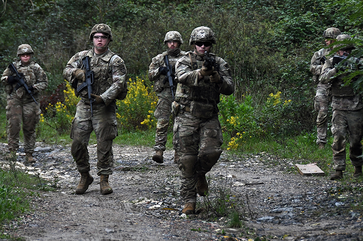A group of six soldiers in helmets walk on a gravel road in a wooded area, their rifles held at the ready.