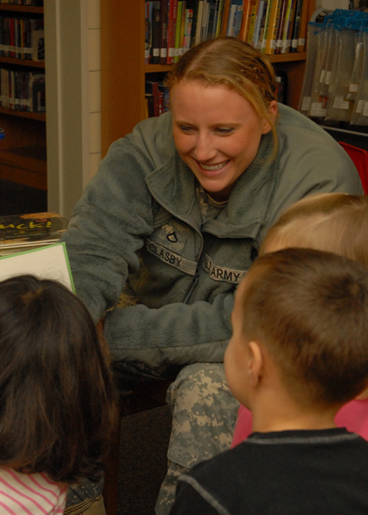 In a tightly framed photograph, a Soldier sits in a library and smiles as she interacts with a group of children. The back of the heads and shoulders of three of them fill the image’s foreground.