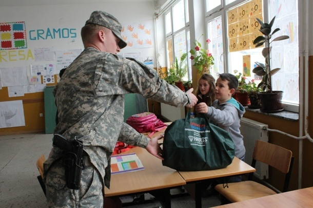 A Soldier in cap and uniform stands in what looks like an elementary school classroom, and he gives a green duffel bag to a boy in a gray hoodie sitting beside a girl at a table.