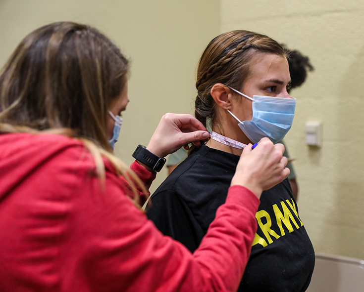 A tightly framed photo shows a woman in a red hoodie placing a white adhesive band to the neck of a second woman, whose blonde hair is gathered in a ponytail and who wears a black T-shirt with “Army” printed in yellow lettering on its front.