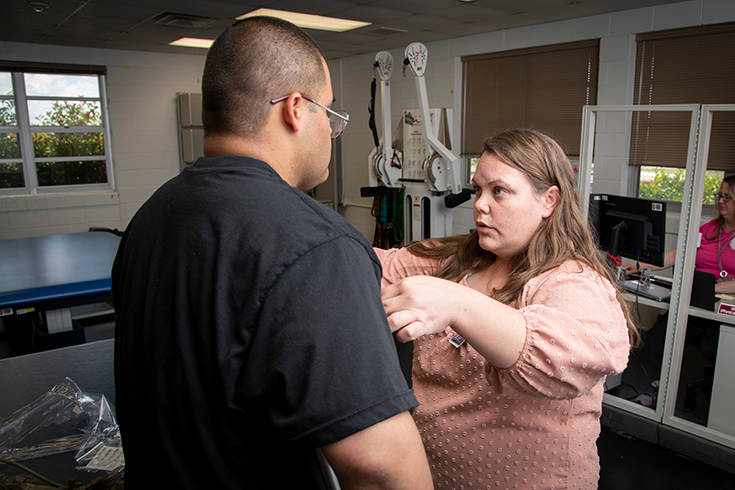 A photograph shows the interior of what looks like a medical setting, with a specialized cot and equipment in the background. In the foreground, a woman in a salmon-colored blouse places her hands on either side of the torso of the man standing before her. She speaks while doing so. The man has a stocky build, a closely shaved head, wire-framed glasses, and a black T-shirt. His back is to the camera, and only a quarter of his profile is visible, making his identity indiscernible.