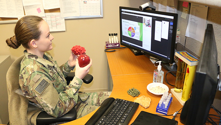 A Soldier with auburn hair gathered in a tight bun at the back of her head sits at a curved wooden desk with two monitors atop it, a keyboard, and a bottle of hand disinfectant. The desk abuts a brown wall, and behind the screens upon it is a gray cubicle wall with papers and sticky notes attached to it. Above one monitor is a tubular camera, about the size of a roll of quarters. The Soldier directs her attention to the device, holding two objects before her as if to demonstrate them to an online audience. One is a shiny red apple, the other a pink, lumpy mass that may be a plastic model of an internal organ, a flower, raw hamburger, or something else.