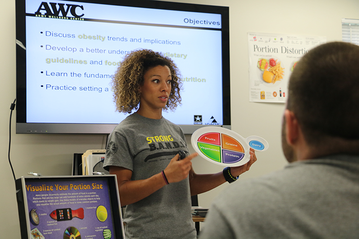 A woman with curly hair stands before a screen mounted on the wall behind her, displaying text with bullet points and an “AWC” logo at its top. The woman holds a circular visual aid before her, with colorful quadrants and white lettering within each pie slice. She gestures toward it while speaking. In the foreground, we see the head and shoulders of a man in a gray T-shirt, suggesting that the woman is presenting to a seated audience before her, beyond the photograph’s frame.
