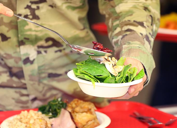 A close-up photo shows a uniformed Soldier filling a paper bowl with a salad.
