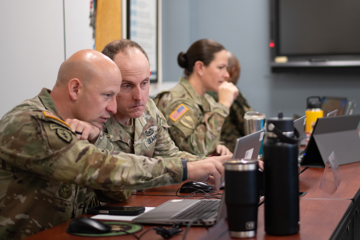 A tightly framed photograph shows a pair of Soldiers sitting at a table. One points to his laptop’s screen, while the other stares at the item indicated. Beyond them, two other Soldiers sit at the table. One seems to stare at her laptop, while the other turns his head to the front of the room, perhaps to look at a whiteboard or instructor beyond the photograph’s frame.