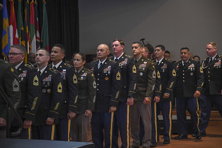 A line of Soldiers in uniform stand in what looks like an auditorium, with long black curtains and a colorful group of flags in the background. The lower left corner of the photograph contains a flat, gray, rectangular surface, suggesting a stage that the men and women are queued up to mount.