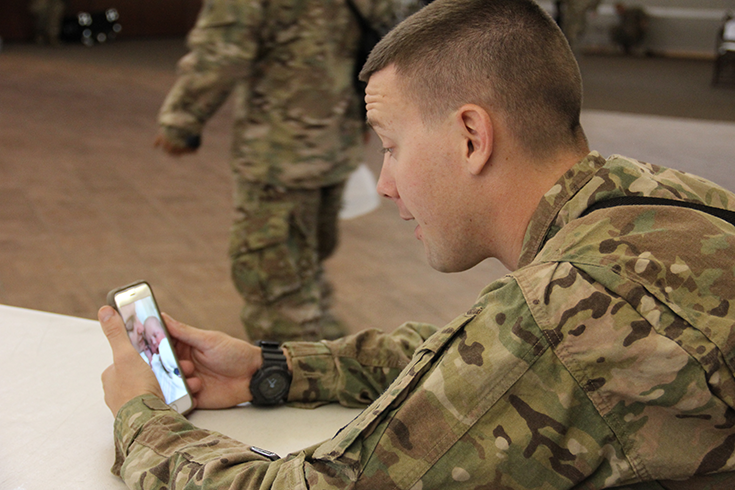 A Soldier sits at a desk, looking at a smartphone whose screen shows an image of a woman holding a baby. In the background of the photograph, beyond the seated man, another Soldier walks past, paper in hand.
