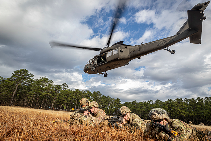 A quartet of helmeted Soldiers lie belly to the ground in a field of brown grass, staring down the sights of the rifles raised before them. A helicopter hovers in the cloudy blue sky above, its blades blurred as it chops the air.