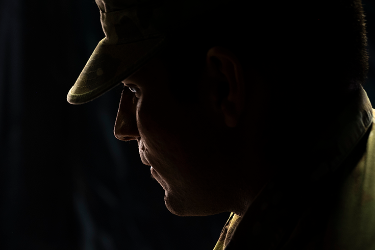 A close-up photograph of a Soldier in a cap shows his profile against a black background. He casts his gaze downward, giving him a pensive look. The image is what photographers describe as low key: lit by a single, soft light source, it reveals only the outlines of the subject’s eyes, nose, chin, and chest, which appear as a white line against the otherwise uninterrupted black obscurity that fills the frame.