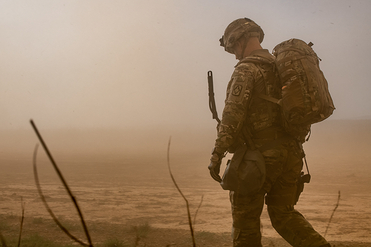 A helmeted Soldier with a pack on his back stares at his boots as he walks along a desolate plain. Dust makes the distant horizon hazy, casting a muted, brownish pall over the whole photograph.