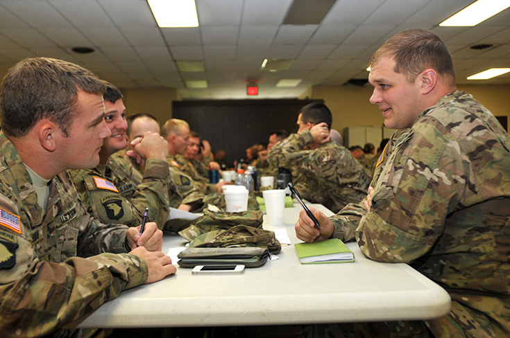 A group of Soldiers sit on either side of a white table inside a room with a drop ceiling and yellow walls. They speak among themselves, some holding pens poised over notebooks. They are bareheaded, and one can see their caps on the table, along with their notebooks, Styrofoam cups, Thermoses, and drink bottles.