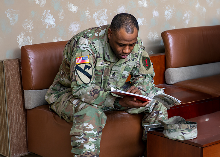 A uniformed Soldier reads a magazine as he sits in what looks like a waiting room, his cap and smartphone placed on the table before him.