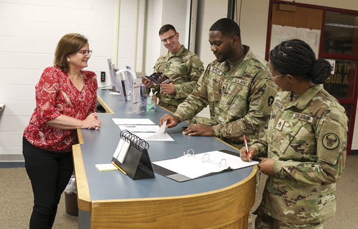 A woman in glasses, a red blouse, and black slacks stands behind a wooden counter with a gray top extending from the foreground of the photograph’s frame to a white cinderblock wall in its background. On the other side of the counter, three uniformed Soldiers stand. One consults a brochure from a stand on the counter, another lifts a page from a group of papers, and the last writes into an open binder. All four smile and seem to interact with one another, their focus turned to the Soldier who turns the pages at the photograph’s center.