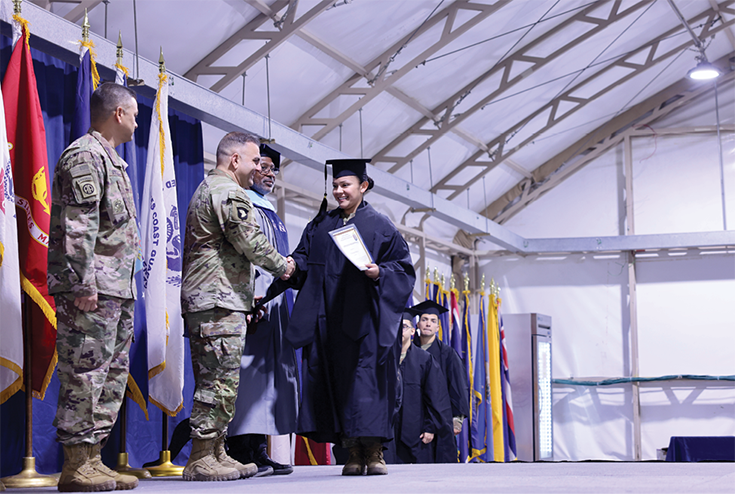 A woman in cap and gown with what resembles a diploma in her left hand uses her right hand to shake the hand of a uniformed Soldier who stands between another Soldier and a man in a professor’s cape and garb. Behind the men are a line of flags, and behind the woman, a line of others in cap and gown. An interior shot, the photograph’s action takes place within a structure with high beams and what appear to be white canvas walls, suggesting a tent-like structure capable of housing military vehicles for ground or air.