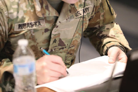 A close-up photograph shows the arms and chest of a Soldier who writes into a white notebook with a teal-colored mechanical pencil.