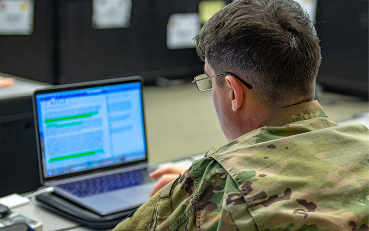 A tightly framed photograph shows the back of the head and shoulders of a bespectacled Soldier who sits at a desk and works on a laptop. Because of the image’s limited depth of field, the Soldier is in focus while his computer’s screen is blurred. However, one can make out a highlighted text document in the onscreen window of an editor such as Microsoft Word.