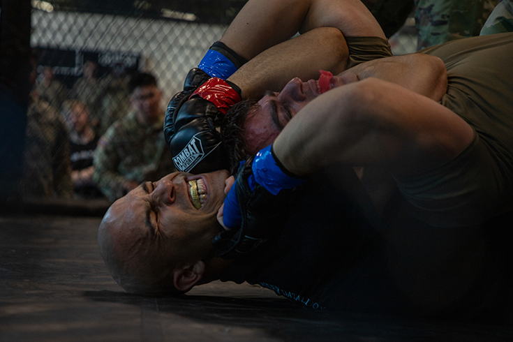A close-up photograph shows the heads and shoulders of a pair of fighters wrestling on a black mat. One fighter, on his back, holds the other to his chest in a chokehold. The other raises his gloved hands toward the head of his adversary. In the background, the blurred figures of Soldiers sit and watch through fencing that defines the edge of the fighting arena.
