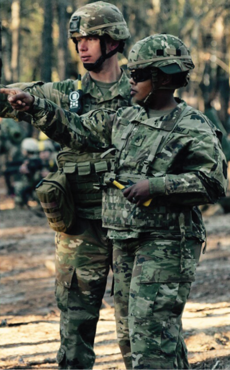 A vertical photograph shows two uniformed Soldiers in a wood, one pointing before them. The gaze of the Soldier leads the viewer’s eye toward the left edge of the photograph. This image is the third in a series of three accompanying the article.